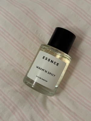 ESSNCE Warm'n Spicy Eau de Parfum - Parfymflaska från ESSNCE med doften Warm'n Spicy. Flaskan är genomskinlig med svart lock och vit etikett med svart text. Modern och stilren design, perfekt för dig som gillar kryddiga och varma doftnoter.