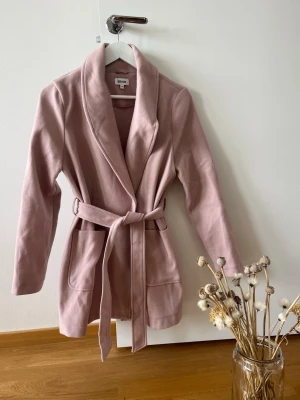 Rosa kappa från Bubbleroom med skärp - Snygg ljusrosa kappa från Bubbleroom med skärp i midjan och stora fickor framtill. Kappan har en klassisk krage och är tillverkad i ett mjukt tyg som ger en stilren look. Perfekt för dig som vill ha en trendig och feminin stil.