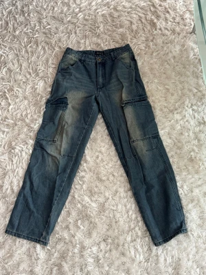 Mörkblå cargobyxor med fickor - Snygga mörkblå cargobyxor i denim med raka ben och flera stora fickor på sidorna. Byxorna har klassisk knapp- och dragkedjestängning samt bakfickor. Perfekta för en avslappnad streetstil.