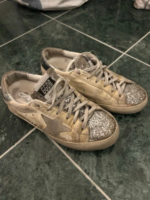 Golden Goose Superstar glitter  - Säljer mina äkta Golden Goose Superstar sneakers i storlek 36. Skick: Använda men i bra skick – slitaget är en del av designen men har även vanligt användningsslitage invändigt (se bilder). Riktigt ovanliga då dom är svåra att få tag på nu.  Storlek: 36 Nypris: ca 5 000 kr Färg: Cream/silver med glitter Material: Läder + mocka Kommer med originalkartong, dustbag och Golden Goose-pass. Hör av dig vid prisförslag eller frågor❤️