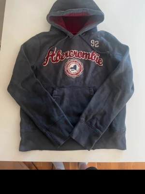 Mörkblå hoodie från Abercrombie - Snygg mörkblå hoodie från Abercrombie & Fitch med röd och vit broderad logga på bröstet och siffran 92 på axeln. Hoodien har en stor huva med rött innerfoder och en klassisk magficka framtill. Perfekt för en avslappnad och cool stil.
