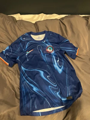 Chelsea FC matchtröja Palmer #20 - Säljer en blå Chelsea FC matchtröja från Nike med Palmer och nummer 20 på ryggen. Tröjan har ett unikt mönster i olika blå nyanser, orange detaljer på ärmsluten och klubbens emblem på bröstet. Perfekt för dig som vill representera laget på läktaren eller träningen.