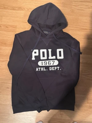 Mörkblå hoodie från Polo Ralph Lauren - Snygg mörkblå hoodie från Polo Ralph Lauren med stor vit logga och texten 'POLO 1967 ATHL. DEPT.' på bröstet. Klassisk huva med dragsko och magficka framtill. Perfekt för en avslappnad och sportig stil.