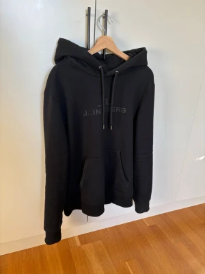  J.Lindeberg hoodie - Svart hoodie från J.Lindeberg ”alpha Hood” med broderad logga på bröstet. Klassisk modell med stor huva, dragsnören och känguruficka framtill. Tillverkad i mjukt material som känns skönt mot huden. Nästintill ny Storlek: L