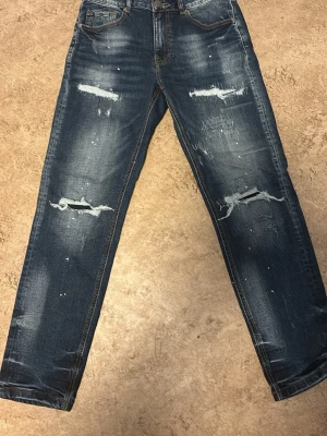Blå slitna jeans med raka ben - Säljer ett par blå jeans med raka ben och tydliga slitningar samt hål på framsidan. Har bara använt dom 1 gång. Jeansen har klassisk femficksmodell och coola detaljer som blekta partier och färgstänk. Perfekta för en avslappnad streetstil.
