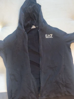Svart hoodie från EA7 Emporio Armani - Svart hoodie med dragkedja från EA7 Emporio Armani. Klassisk design med huva och logga på bröstet. Perfekt för en avslappnad och sportig stil. Mjuk bomullskänsla och fickor på sidorna.