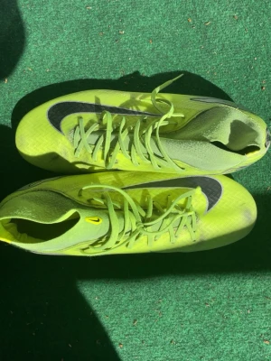 Nike Mercurial gröna fotbollsskor - Säljer ett par Nike Mercurial fotbollsskor i neon-grönt med svarta detaljer och silverfärgade inslag. Skorna har snörning och dobbar för spel på gräs. Lätta och smidiga med en modern, strömlinjeformad design som passar dig som vill ha fart på planen. Storlek 45 och en av snören är av 