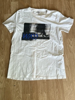 Vit t-shirt med tryck från WARP - Vit t-shirt från WARP med svartvitt grafiskt tryck och blå text på bröstet. Klassisk rund hals och korta ärmar. Tillverkad i mjuk bomull för skön känsla och enkel stil.