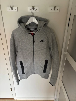 Grå Nike tech fleece hoodie - Snygg grå hoodie från Nike med svart logga på bröstet och hel dragkedja framtill. Två svarta dragkedjeförsedda fickor och huva. Tillverkad i mjukt sweatshirtmaterial som är perfekt för chill eller träning.