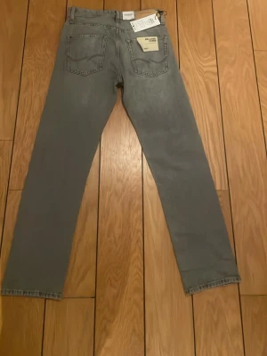 Grå raka jeans från Jack & Jones - Säljer ett par grå jeans från Jack & Jones med rak passform och klassisk femficksdesign. Jeansen har knappgylf och är tillverkade i ett slitstarkt bomullsmaterial. Perfekta för en clean och avslappnad stil. Storlek W27 L32