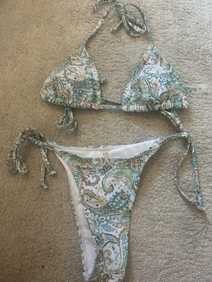  OANVÄND Mönstrad bikini med knyt i XS - Snygg bikini med paisleyinspirerat mönster i vitt, grönt och guld. Överdelen är trekantsmodell med knyt i nacken och ryggen, underdelen har knyt i sidorna. Tillverkad i mjuk polyester och elastan, perfekt för stranddagar.