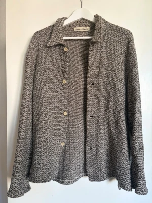 Gråmelerad overshirt från Our Legacy - Säljer en gråmelerad overshirt från Our Legacy med avslappnad passform och klassisk krage. Skjortan har synliga knappar framtill och är tillverkad i ett grovt, stickat material som ger en unik struktur. Perfekt lager på lager-plagg för dig som gillar stilrena detaljer. 100% bomull/cotton