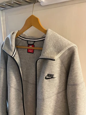 Nike tech  - Snygg grå hoodie från Nike med svart logga på bröstet och dragkedja framtill. Hoodien har svarta detaljer längs dragkedjan och runt huvan. Tillverkad i mjukt material som passar perfekt för en sportig och avslappnad stil.