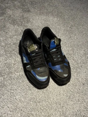 Valentino rockrunner blå camo sneakers - Snygga sneakers från Valentino med blå, svarta och grå kamouflagemönster. Skorna har svarta snören, svart sula och detaljer i mocka och textil. Perfekta för dig som vill sticka ut med en unik och trendig stil.