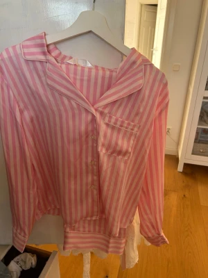 Rosa- och vitrandig pyjamas set - Säljer en söt pyjamasset från Primark i storlek XS. Den har rosa och vita vertikala ränder, klassisk krage, knappstängning framtill och en bröstficka. Materialet är mjukt och lent, perfekt för mysiga kvällar hemma.