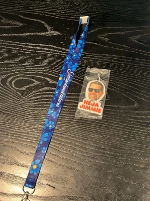 Blått nyckelband med blommor - Säljer ett blått nyckelband med blommönster och text, samt en nyckelring med texten 'Heja Jimmie' i vitt och rött. Perfekt för att hålla koll på nycklar eller accessoarer.