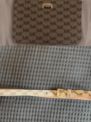 Beige MK plånbok med logomönster + matchande skärp - Snygg beige plånbok från Michael Kors med klassiskt MK-logomönster över hela ytan. Plånboken har guldfärgade detaljer och dragkedja upptill. Matchande skärp i samma mönster och färg med guldfärgat spänne. Perfekt accessoar för dig som gillar stilrena detaljer.