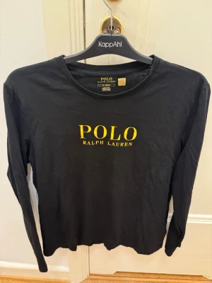 Svart långärmad pyjamastopp Polo - Svart långärmad pyjamastopp från Polo Ralph Lauren i mjuk bomull. Tröjan har rund halsringning och gul logotyp med text på bröstet. Enkel och stilren design som passar perfekt för avslappnade kvällar.