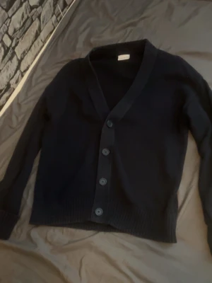 Mörkblå cardigan från John Henric - Klassisk mörkblå cardigan från John Henric med v-ringning och fyra knappar framtill. Tröjan har långa ärmar och ribbade muddar vid ärmslut och nederkant. Perfekt att slänga över en t-shirt eller skjorta för en clean look.