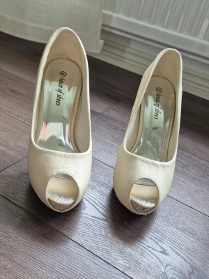 Vita peep toe pumps från House of Shoes - Säljer ett par eleganta vita pumps från House of Shoes med peep toe och hög klack. Skorna har en klassisk siluett och är tillverkade i tygmaterial med glansig innersula. Perfekta för dig som vill ha en stilren och chic look. 12cm klack