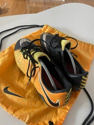 Nike Hypervenom fotbollsskor - Säljer ett par Nike Hypervenom fotbollsskor i svart och gul färg med vita detaljer. Skorna har snörning, låg profil och mönstrad ovandel för bättre bollkontroll. Sulan har fasta dobbar för spel på gräs. Medföljer en matchande gul Nike-påse.