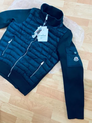Svart quiltad jacka från Moncler - Snygg svart quiltad jacka från Moncler med dragkedja framtill och två fickor med dragkedja. Jackan har hög krage, långärmade ärmar och en Moncler-logga på ärmen. Perfekt för dig som vill ha en stilren och modern look.