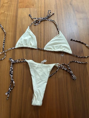 KrämVit bikini med leopardmönstrade band - Snygg vit bikini med trekantsöverdel och knyt i sidorna. Banden har ett coolt leopardmönster som ger en trendig touch. Tillverkad i mjuk polyesterblandning, perfekt för strand eller poolhäng i sommar. Helt oanvänd.
