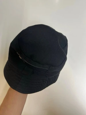Svart bucket hat med ficka - Säljer en svart bucket hat i tyg med en liten ficka med dragkedja på sidan. Hatten har klassisk form med mjuk brätte och är perfekt för en avslappnad streetwear-look. Materialet känns slitstarkt och stilen är enkel men cool.