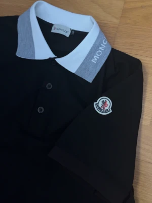Svart Moncler pikétröja med logga - Svart pikétröja från Moncler med gråvit krage och vit Moncler-text på kragen. Klassisk knappslå med tre knappar och broderad Moncler-logga på ärmen. Perfekt för en clean och stilren look.