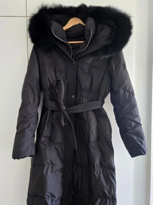 Svart dunjacka med pälshuva - Svart lång dunjacka med bälte och stor huva med fluffig päls. Silverfärgad dragkedja och knappar framtill samt ribbade detaljer vid bältet och huvan. Perfekt för kalla vinterdagar och är riktigt varm. Köptes ht 2024/vt 2025 och användes samma vinter. Därefter har den varit oanvänd då jag köpte en ny vinterjacka. Älskade modellen men ångrade köpet då jag inte föredrar silverddetaljer. 