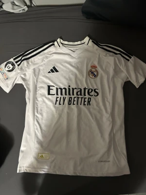 Real Madrid matchtröja Adidas vit - Vit Real Madrid matchtröja från Adidas med svarta detaljer och klubbmärke på bröstet. Tröjan har trycket 'Emirates Fly Better' och svarta ränder på axlarna. På ärmen finns Foundation-märke och Champions League-märke med siffran 15. Material: polyester.