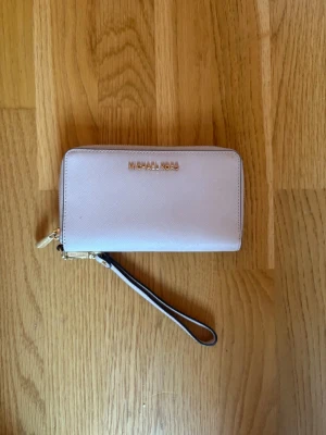 Ljusrosa plånbok Michael Kors - Snygg ljusrosa plånbok från Michael Kors i ett tåligt saffianoläder. Plånboken har guldfärgade detaljer, dragkedja runtom och en avtagbar handledsrem. Perfekt för dig som vill ha både stil och funktion i ett.