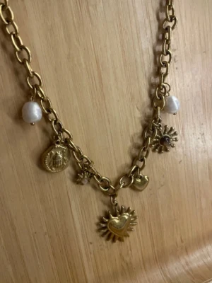 Guldigt halsband med berlocker och pärlor - Unikt halsband i guldfärgad metall med chunky kedja och flera berlocker, inklusive solformad medaljong, hjärta, mynt och vita oregelbundna pärlor. Perfekt för dig som gillar statement-smycken och vill sticka ut med coola detaljer.