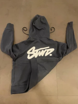 Mörkgrå SWD hoodie med tryck - Snygg mörkgrå hoodie från SWD med stor vit logga i graffiti-stil på ryggen. Klassisk huva och känguruficka framtill. Tillverkad i mjukt bomullsmaterial som är perfekt för chill dagar. Oversized fit för en avslappnad look.