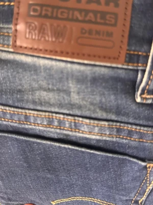 Blå jeans från G-Star RAW - Säljer ett par klassiska blå jeans från G-Star RAW med snygga kontrastsömmar och dragkedjegylf. Jeansen har en patch med logga bak i midjan och detaljerade nitar. Tillverkade i slitstarkt denim, perfekta för en cool och avslappnad stil.