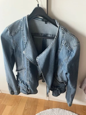 Blå jeansjacka med detaljer - Snygg blå jeansjacka från Kahpal med markerade axeldetaljer och dekorativa sömmar. Jackan har bred krage, dragkedjor vid fickorna och volangliknande kant nertill. Perfekt för dig som vill ha en unik twist på klassisk jeansjacka. Stresch passar 42 och mer