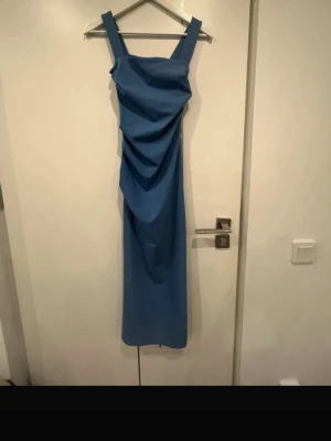 Blå långklänning - Snygg blå långklänning med breda axelband och draperad detalj framtill. Klänningen har en rak passform och är ärmlös, vilket ger en stilren och modern look. Perfekt för dig som vill sticka ut med en enkel men elegant stil.