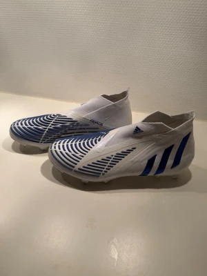 Adidas Predator vita/blå fotbollsskor - Säljer ett par Adidas Predator fotbollsskor i vitt med blåa detaljer och mönster. Skorna har en strumpliknande passform utan snörning och är tillverkade i syntetmaterial för bästa bollkontroll. Perfekta för dig som vill sticka ut på planen.