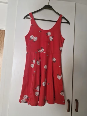 Röd blommig sommarklänning - Söt röd kortklänning med vita blommor och gröna blad. Klänningen har ärmlös design, rundad halsringning och utställd kjol. Perfekt för varma dagar och enkel att styla med sneakers eller sandaler.