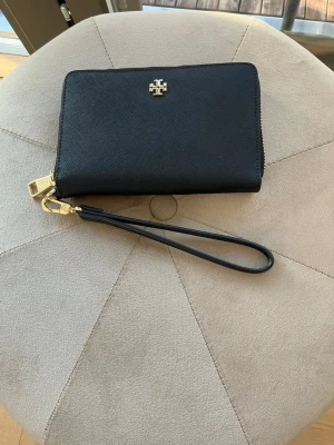 Svart plånbok från Tory Burch - Snygg svart plånbok från Tory Burch i strukturerat läder med guldfärgade detaljer och avtagbar handledsrem. Plånboken har dragkedja runtom och den ikoniska loggan framtill. Går att använda som en liten väska. Tillräckligt stor för att rymma det nödvändigaste som vanlig storlek på mobiltelefon, kort, pengar och nycklar.  Väl omhändertagen med endast sparsamma tecken på öppning och stängning vid dragkedjan som enda anmärkning. 