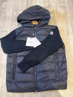 Svart pufferjacka från Moncler - Snygg svart pufferjacka från Moncler med stickade ärmar och klassisk huva. Jackan har dragkedja framtill, två fickor med dragkedja och Moncler-logga på ena ärmen. Perfekt för kalla dagar och riktigt trendig look.