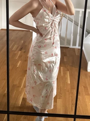 Ljusblommig långklänning med snörning - Söt långklänning i ljus ton med små rosa blommor, tunna axelband och snörning framtill som ger en snygg detalj. Öppen rygg och figurnära passform. 