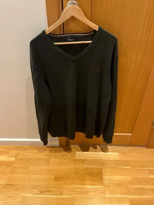 Mörkgrön stickad tröja Fred Perry - Mörkgrön stickad tröja från Fred Perry med V-ringning och diskret broderad logga på bröstet. Klassisk och enkel design med ribbade muddar vid ärmslut och nederkant. Perfekt för dig som gillar stilrena plagg.
