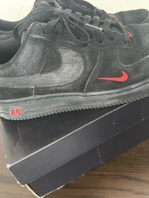 Nike Air Force 1 svart/röd suede - Snygga Nike Air Force 1 sneakers i svart mocka med röda detaljer på Swoosh och AIR-loggan. Skon har en unik grå mönstrad Swoosh på sidan och klassisk låg siluett. Perfekt för dig som vill sticka ut med stilrena och coola sneakers.