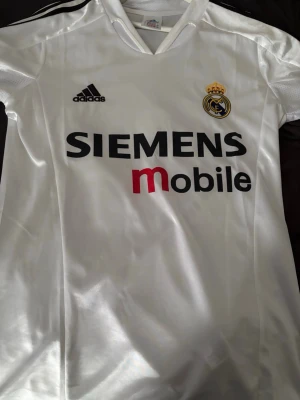 Real Madrid matchtröja Zidane #5 - Säljer en vit Real Madrid matchtröja från Adidas med svarta detaljer och klubbens emblem på bröstet. Tryck med SIEMENS mobile på framsidan och Zidane #5 på ryggen. 