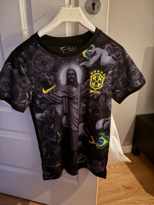 Svart Brasilien fotbollströja Nike - Snygg svart fotbollströja från Nike med Brasilien-tryck och detaljer i gult och blått. Framsidan har ett stort motiv av Kristusstatyn och brasilianska flaggan, samt Brasilien-emblem på bröstet. Tillverkad i lätt och ventilerande material, perfekt för träning eller match.