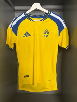 Sverige matchtröja Adidas gul/blå - Gul matchtröja från Adidas för Sveriges landslag med blå och vita detaljer på axlarna. Broderat landslagsmärke på bröstet och Adidas-logga. Tillverkad i lätt och ventilerande funktionsmaterial, perfekt för fotbollsträning eller match. Storlek s passar xs