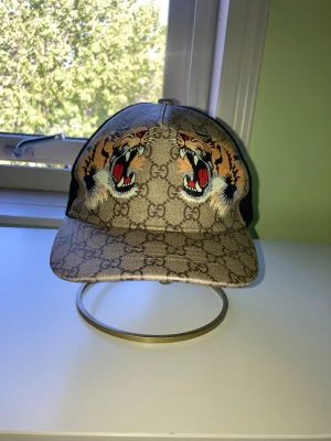 Gucci keps med tigerbrodyr - Snygg keps från Gucci med klassiskt GG-mönster i beige och brunt. Framtill finns två broderade tigrar med starka färger. Kepsen har svart mesh på baksidan och justerbar rem med röd-gröna detaljer. Perfekt för dig som vill sticka ut. SNABB AFFÄR! PRINS KAN SÄNKAS! SKICKA OM NI VILL HA MER BILDER!