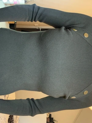 Mörkgrön ribbad topp med knappar - Snygg mörkgrön ribbad topp med lång ärm och dekorativa guldfärgade knappar vid axlarna. Modellen är croppad och har en tight passform som framhäver figuren. Perfekt till jeans eller kjol för en trendig look. inga defekter 