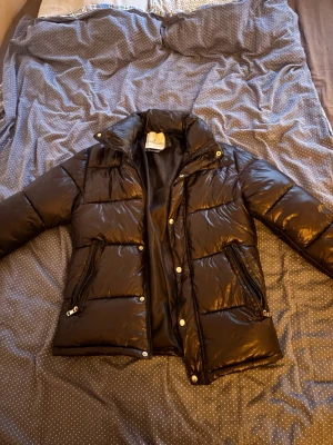 Svart dunjacka från Moncler - Snygg svart dunjacka från Moncler med glansig finish och klassisk logotyp på ärmen. Jackan har dragkedjor på fickorna, hög krage och tryckknappar framtill. Perfekt för kalla vinterdagar och riktigt trendig look.